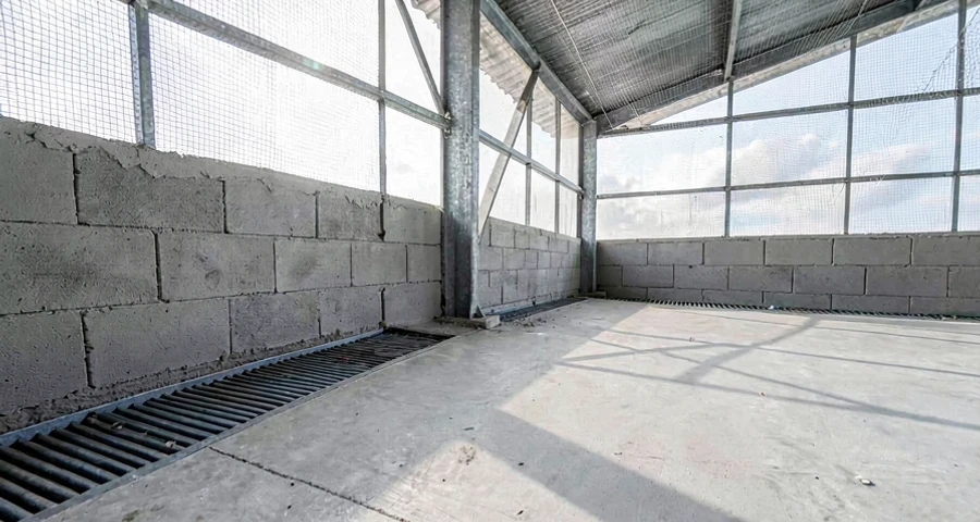 Vista interna de aviário industrial de frango de corte em construção no sul do Brasil, mostrando estrutura metálica galvanizada, contrapiso de concreto recém executado com malha de aço, muretas laterais de blocos e cortinas plásticas amarelas abertas, iluminação natural intensa, operários com EPIs trabalhando, ambiente de obra real